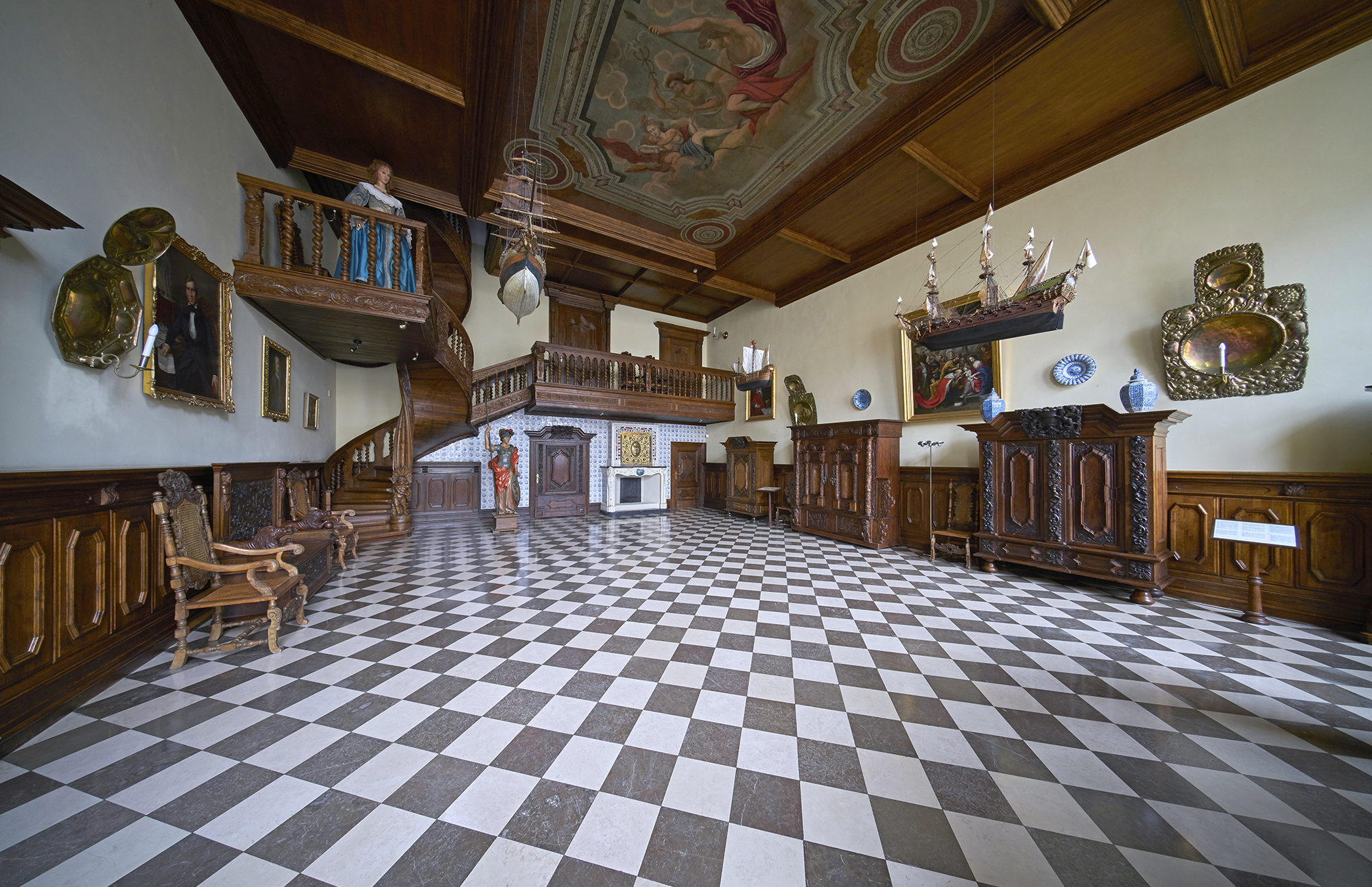 Wnętrze Sieni Gdańskiej w Nowym Domu Ławy. Ściany do połowy wyłożone boazerią, pod nimi stoją meble gdańskie, szafy i krzesła. W głębi drewniana klatka schodowa.